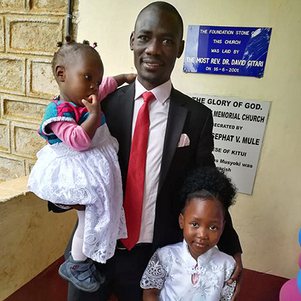 Eric Omondi smiling at the camera. He is holding one of his daughter in his right arm, while his left hand is on his other daughter's arm. They are all dressed elegantly.
